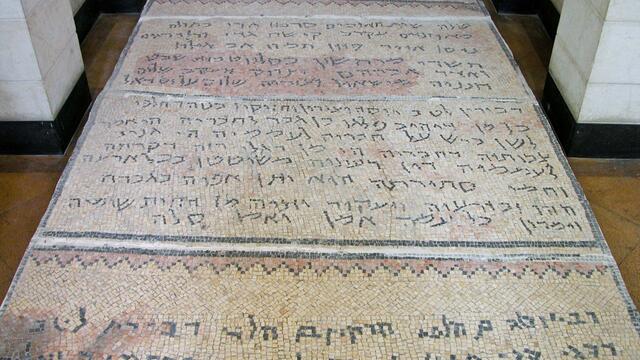 A rectangular floor mosaic with Hebrew and Aramaic inscriptions in black letters, bordered by a geometric pattern, set in a tiled hallway with stone walls.