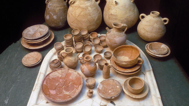 A collection of ancient ceramic jugs, bowls, plates, and cups in various sizes and earthy tones, arranged neatly on a stone surface with several large jugs in the background.