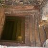 Photo of a rectangular stone pool, partially filled with water, with steps leading down into it, weathered brick and stone walls, and and stairs leading up through a doorway.