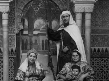 Photo of a family in ornate dress, including a man standing with a staff and two women, seated, one holding a baby, all in front of a building with ornate arches and decorations.