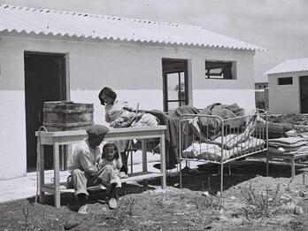 A man and two children sit outside a small building with a corrugated roof, surrounded by worn furniture and bedding.