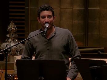Thumbnail photo of man on synagogue bimah speaking into a microphone, with music stands in the foreground and a bass player and Torah crown in the background.