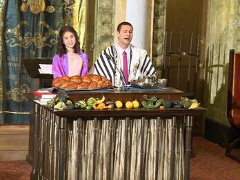 Thumbnail photo of singing man in a suit and girl in a dress, both wearing a tallis, on a synagogue bimah with challah and vegetables on the table in front of them.
