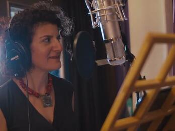 Thumbnail photo of woman wearing headphones and facing a recording microphone, with a music stand in the foreground.