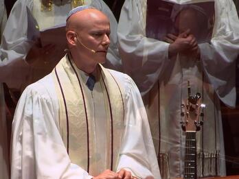 Thumbnail photo of man in white kippa and robes wearing a headset microphone, with several others in the background behind him.