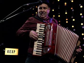 Thumbnail photo of man playing accordion, with decorative lights and "KEXP" lit-up sign in the background.