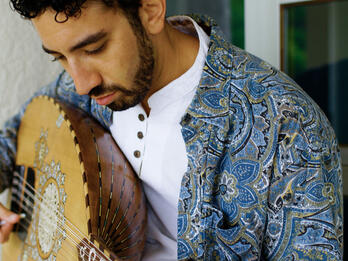 A man in a blue paisley shirt plays an oud.