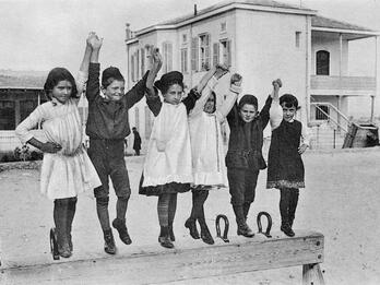 Girls and boys holding hands and jumping on a pommel horse in front of a building.