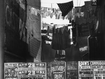 Street view photograph depicting clotheslines above street level, advertisement posters plastered on the wall, and three sets of people walking down the street.