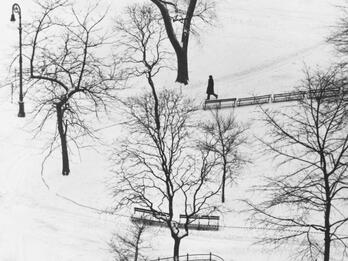 Aerial photograph of a city square covered in snow featuring trees, benches, lampposts, and a man walking through the square.