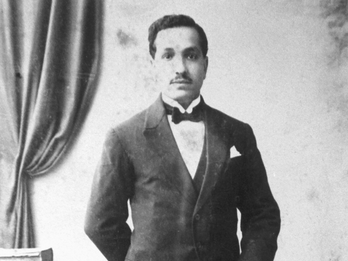 Photograph of man in suit with one arm on a walking stick. 