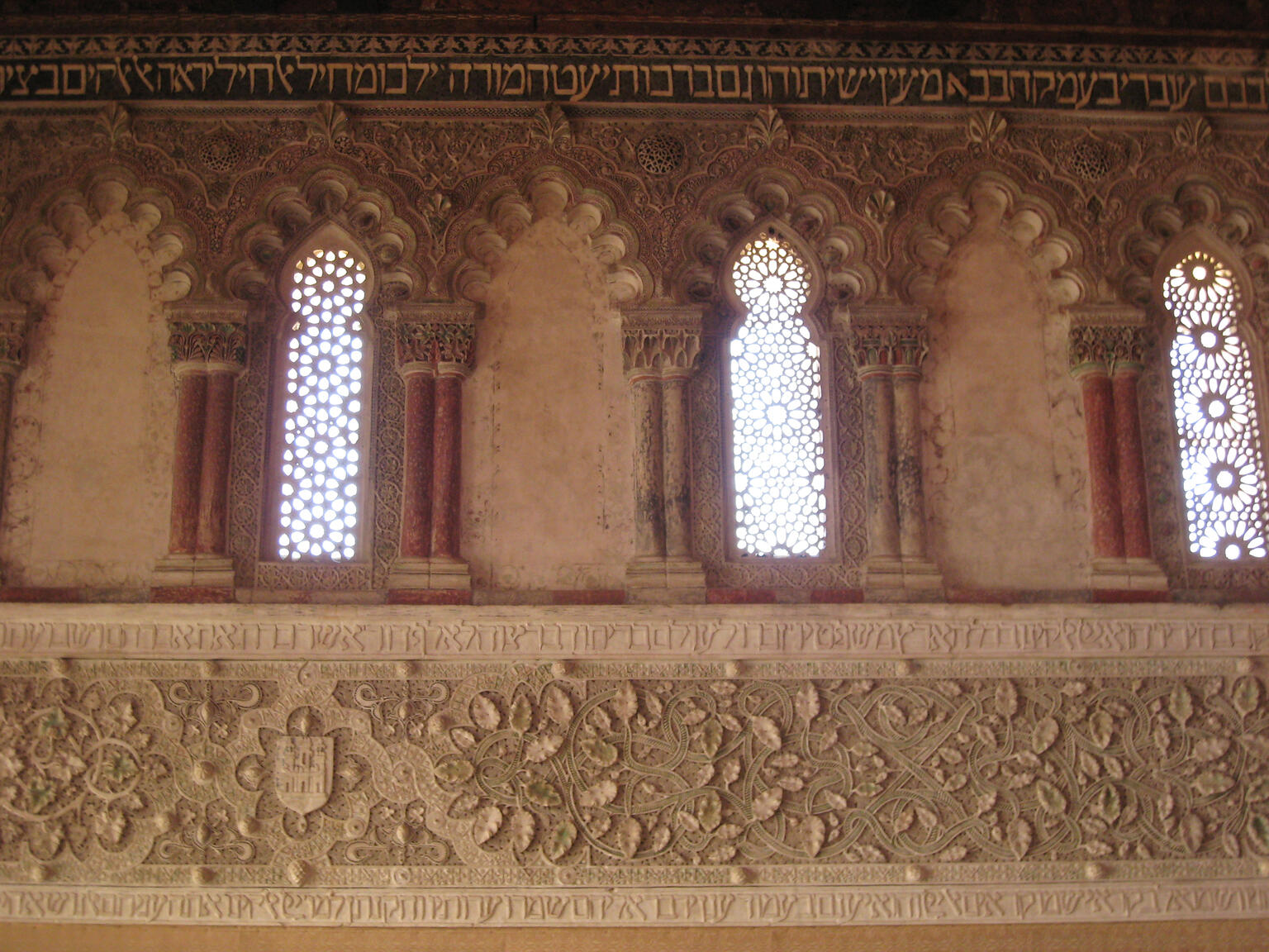 Photo of a wall with a row of ornate arches and windows and a row of ornate floral decorative engraving, each surrounded by three lines of engraved Hebrew text.