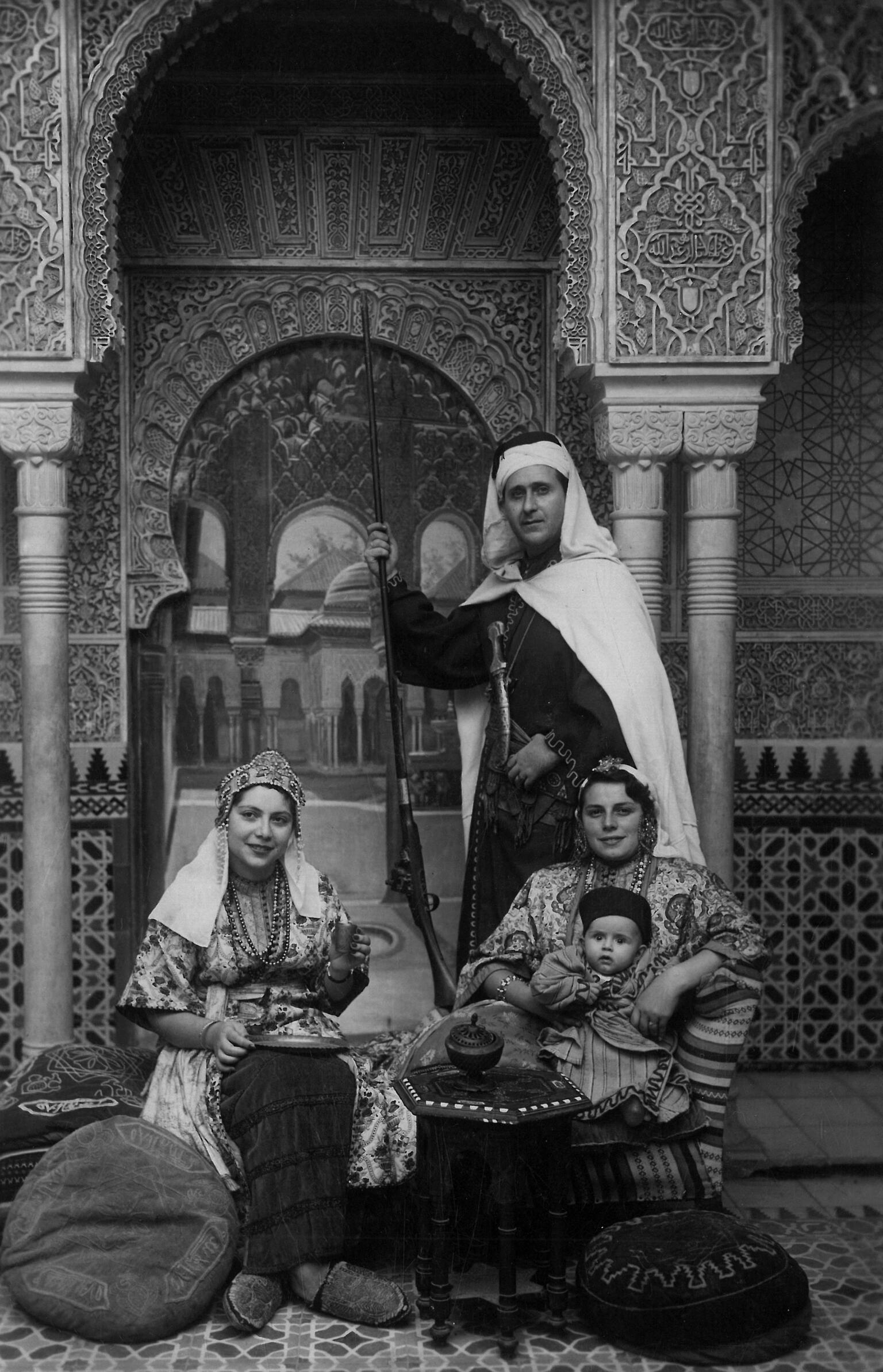 Photo of a family in ornate dress, including a man standing with a staff and two women, seated, one holding a baby, all in front of a building with ornate arches and decorations.