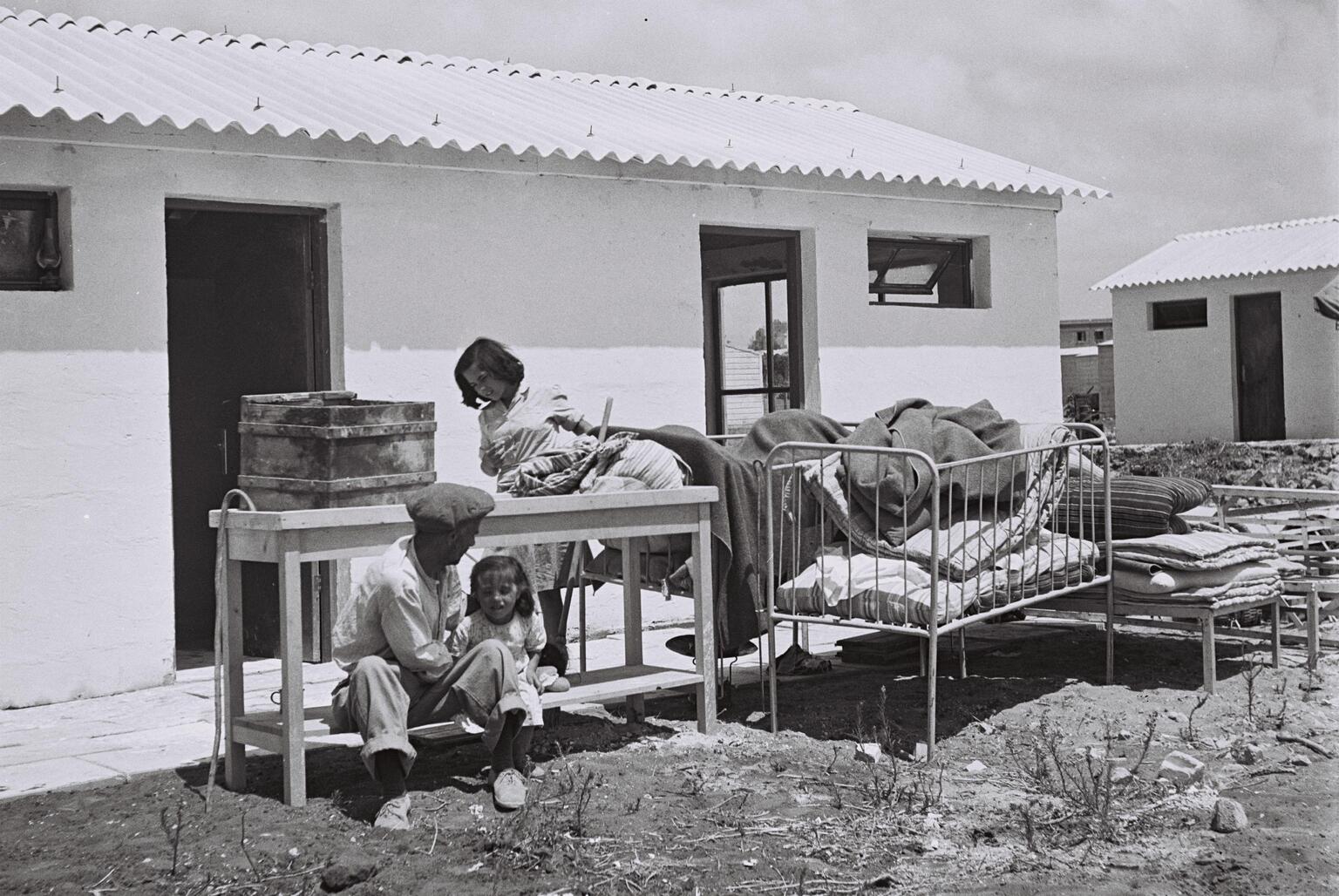 A man and two children sit outside a small building with a corrugated roof, surrounded by worn furniture and bedding.