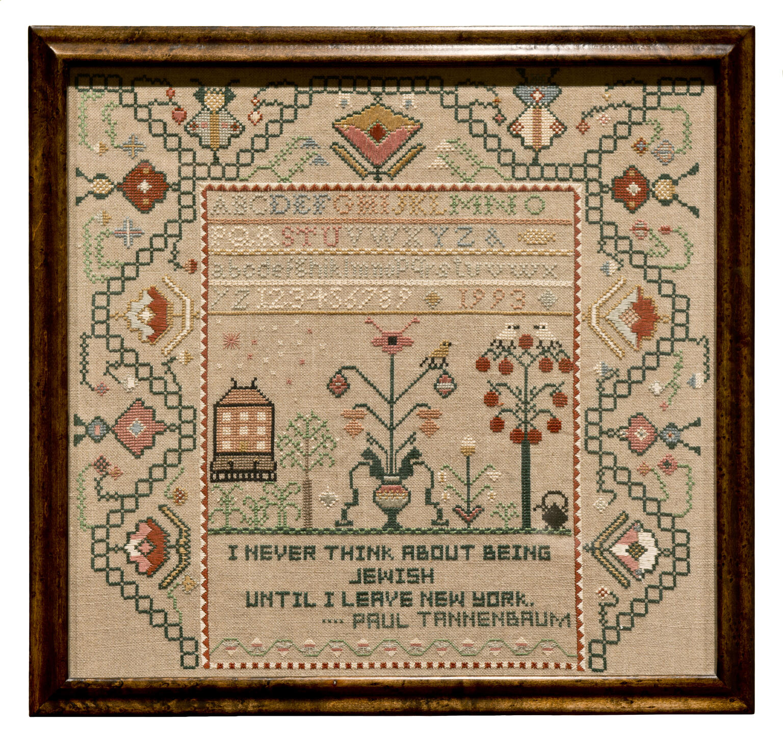 Needlework in a frame with a border of designs, the alphabet and numbers in the center, and text at the bottom.
