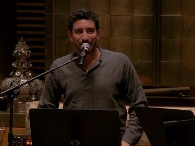 Thumbnail photo of man on synagogue bimah speaking into a microphone, with music stands in the foreground and a bass player and Torah crown in the background.