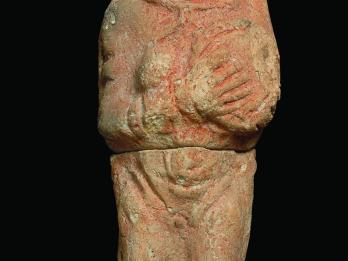 Terra-cotta figurine of woman wearing accessories holding a disk over left breast while right breast is exposed. 