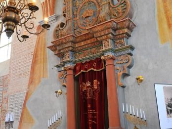 Photograph of carved doorway set in wall with curtain across it, surrounded by candelabra, Hebrew writing on wall, and chandelier in forefront. 