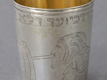 Silver cup engraved with a person in profile wearing a jester hat drinking out of a barrel. 