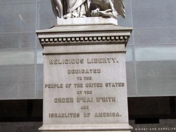 Marble monument depicting a figure dressed in coat and mantle with shield with American flag in relief on breast, with a naked boy under her right arm holding a lamp, and an eagle. 