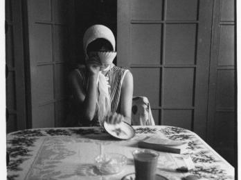 Photograph of woman sitting at the end of a table in front of open doors, wearing a scarf and covering her face with tea cup.