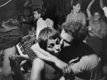 Photograph of several people socializing, including someone with accordian, three figures dancing, and a man with his arm around a woman and kissing her cheek in the foreground.