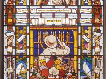 Stained glass window featuring a man in the center facing away from viewer lighting a menorah behind a table set for Shabbat, a rabbi above him next to a harp, and biblical images and Hebrew words, flowers, birds, and other objects.