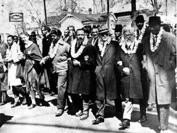 Photograph of men marching down the street with arms linked wearing white double carnation leis. 