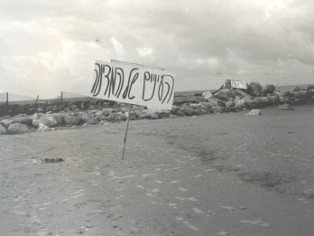 Photograph of Hebrew sign on empty beach with a rock jetty in background.