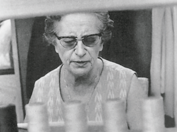Photograph of elderly woman with glasses framed by spools of thread. 