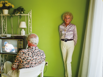 Photograph of older couple in apartment. The woman has bold eye makeup and a perm and stands facing the camera while the man sits in a chair in front of the television watching a baseball game with his back to the camera.