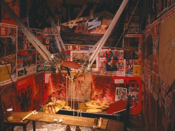 Installation featuring catapult-like contraption in the middle strung between two walls, a hole in the ceiling, and scientific drawings and posters on the walls. 