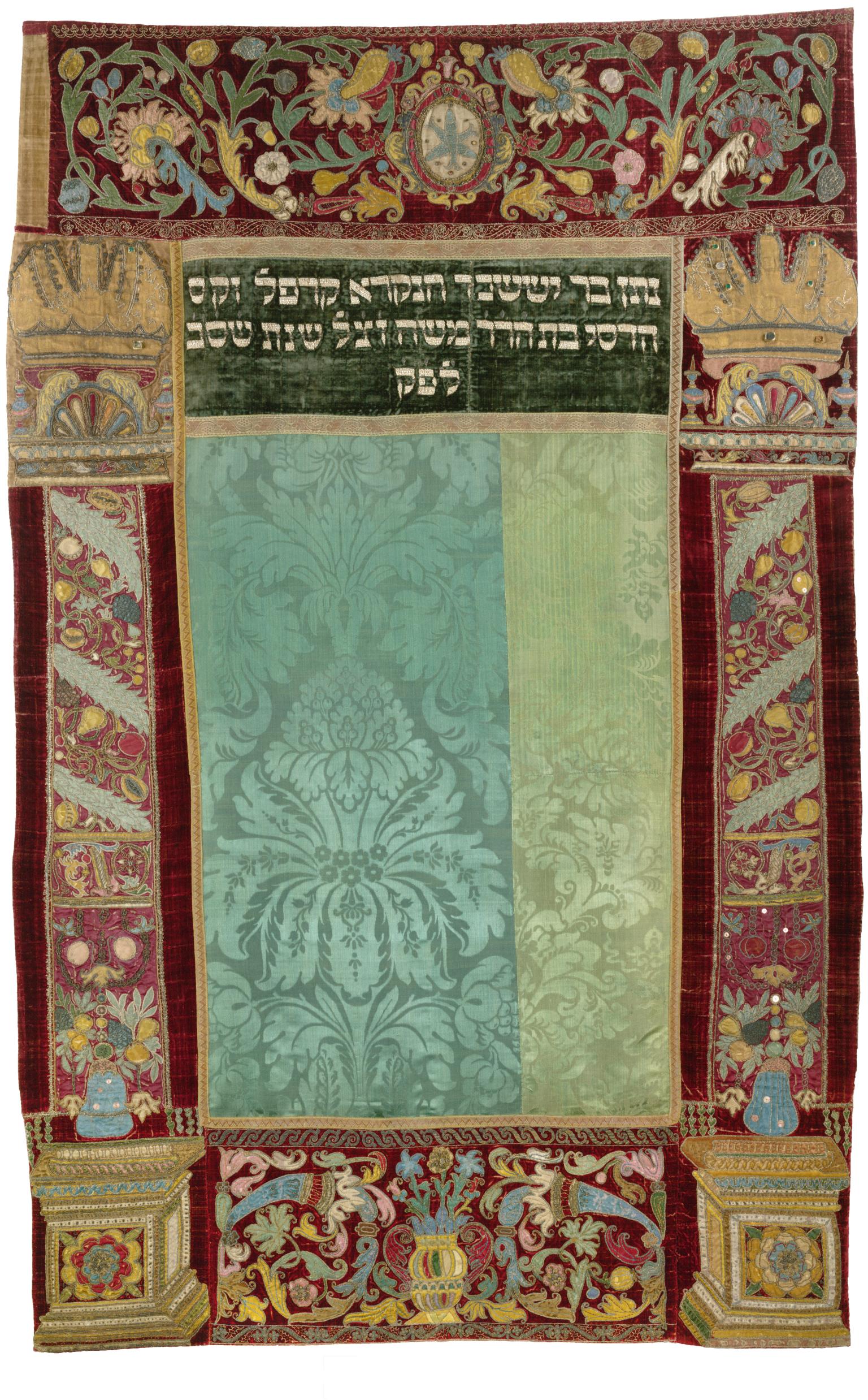 Embroidered curtain of columns with crowns on left and right sides and decorative floral motifs throughout, and Hebrew text in top center.