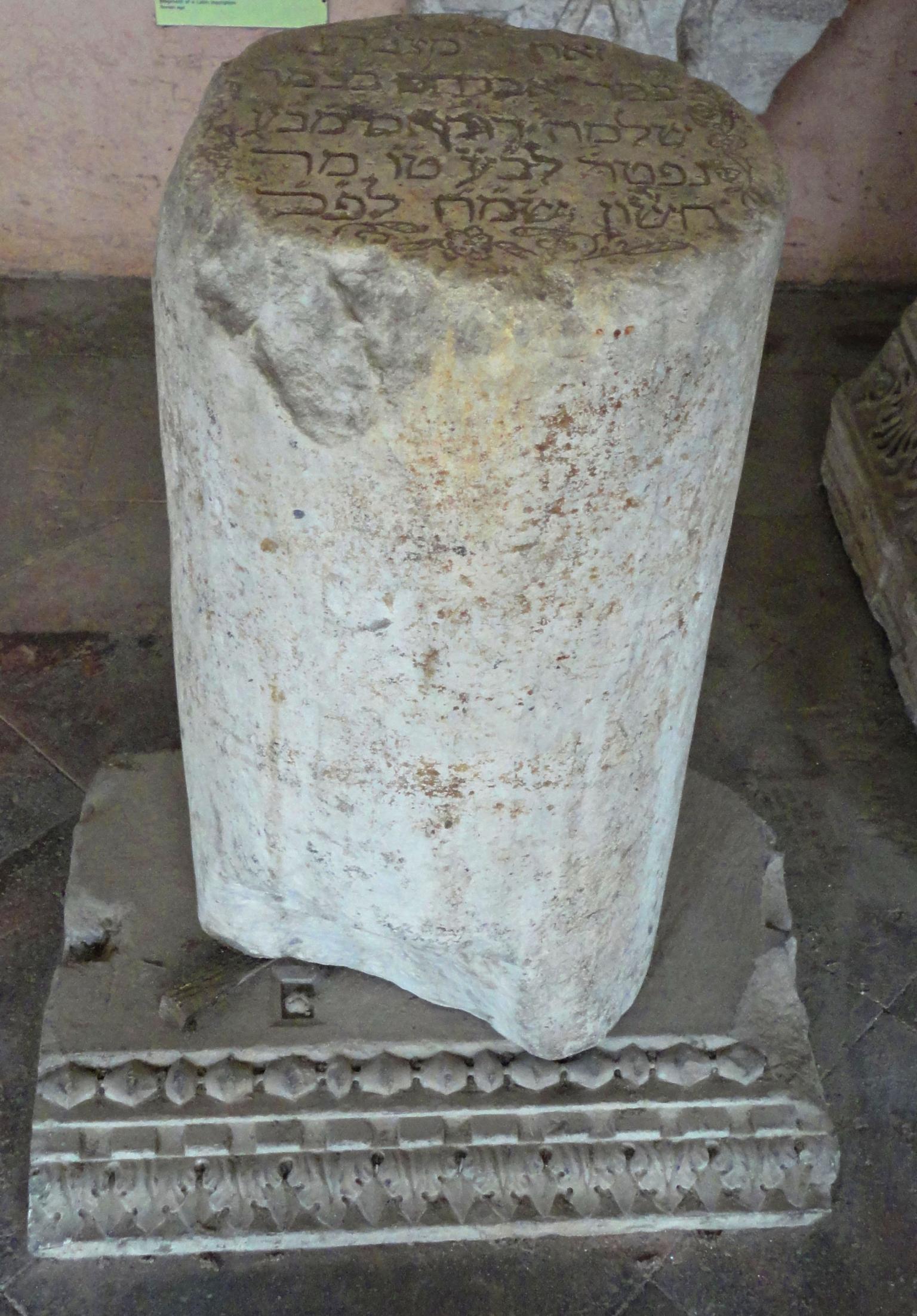 Cylindrical tombstone with Hebrew on top, sitting on decorative base.