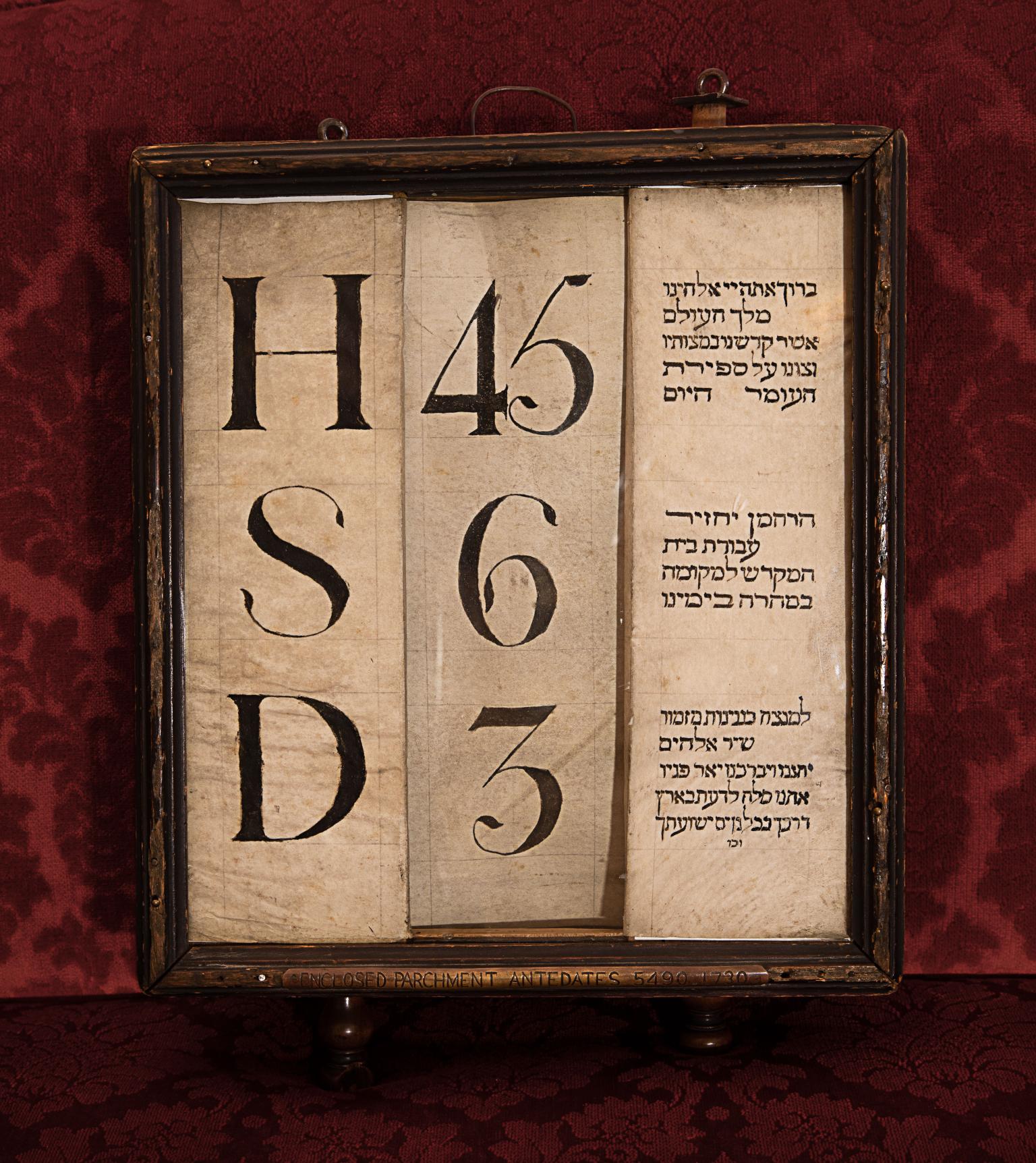 Framed piece of paper with three columns of text: single letters in left column, numbers in middle column, and Hebrew text in right column. 