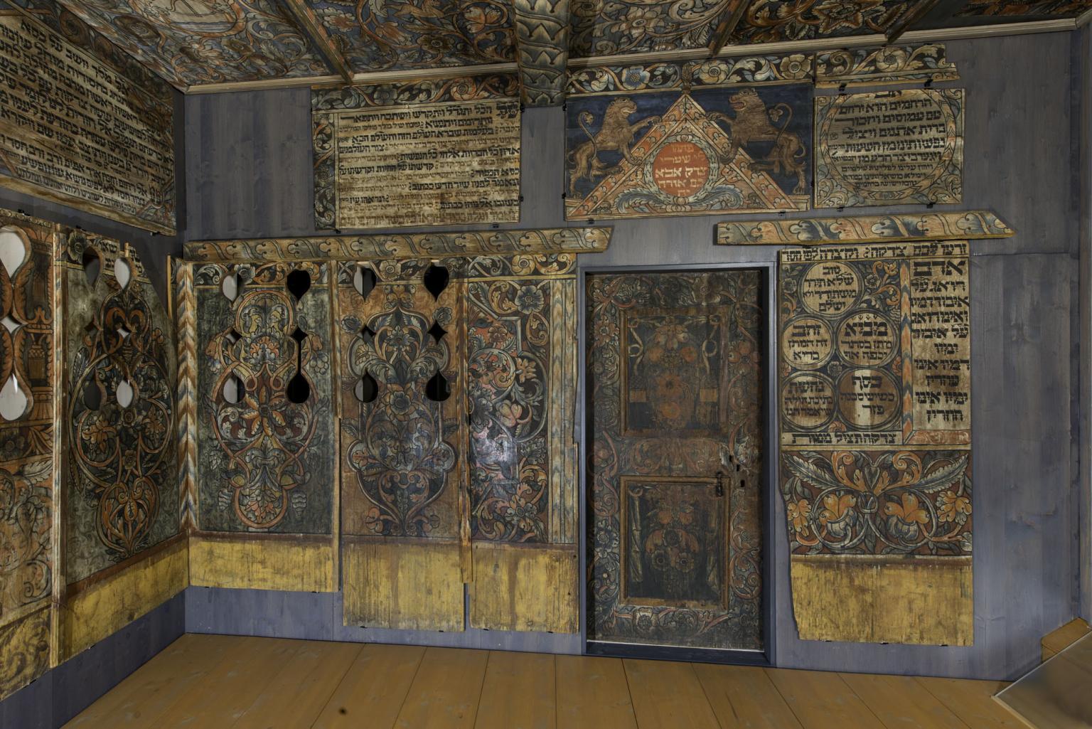 Photograph of room with wood walls painted with Hebrew text, animals, and floral designs.