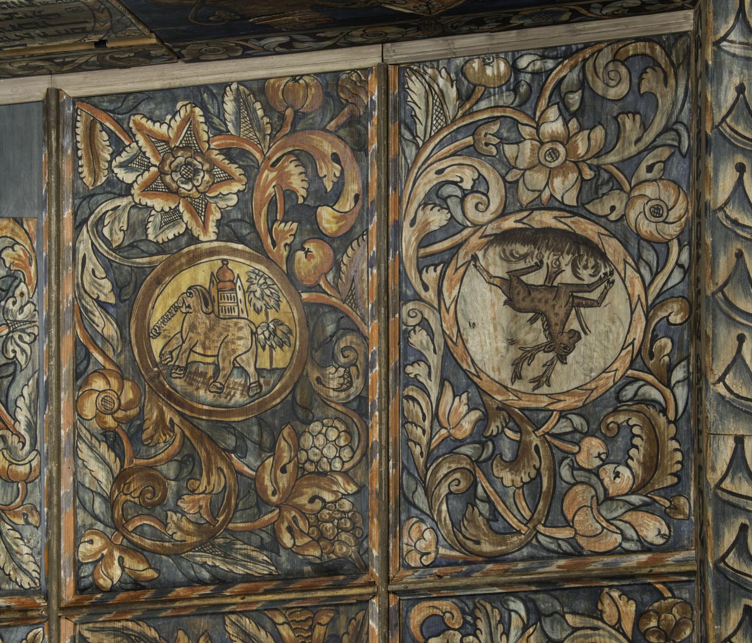 Close-up photograph of wood wall painted with foliage and image of elephant carrying castle and upside-down deer.
