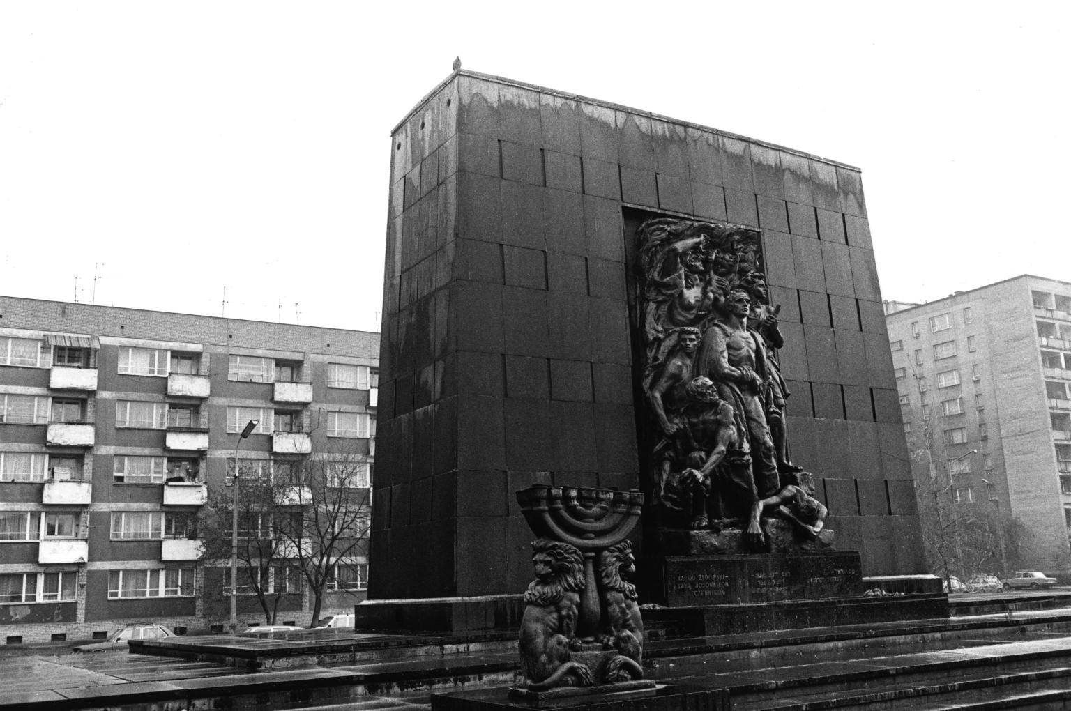 Photograph of tall, rectangular monument in front of apartment buildings, depicting seven figures gathered around central figure and smaller sculpture of a seven-branched menorah flanked by lions in front of monument in left foreground. 