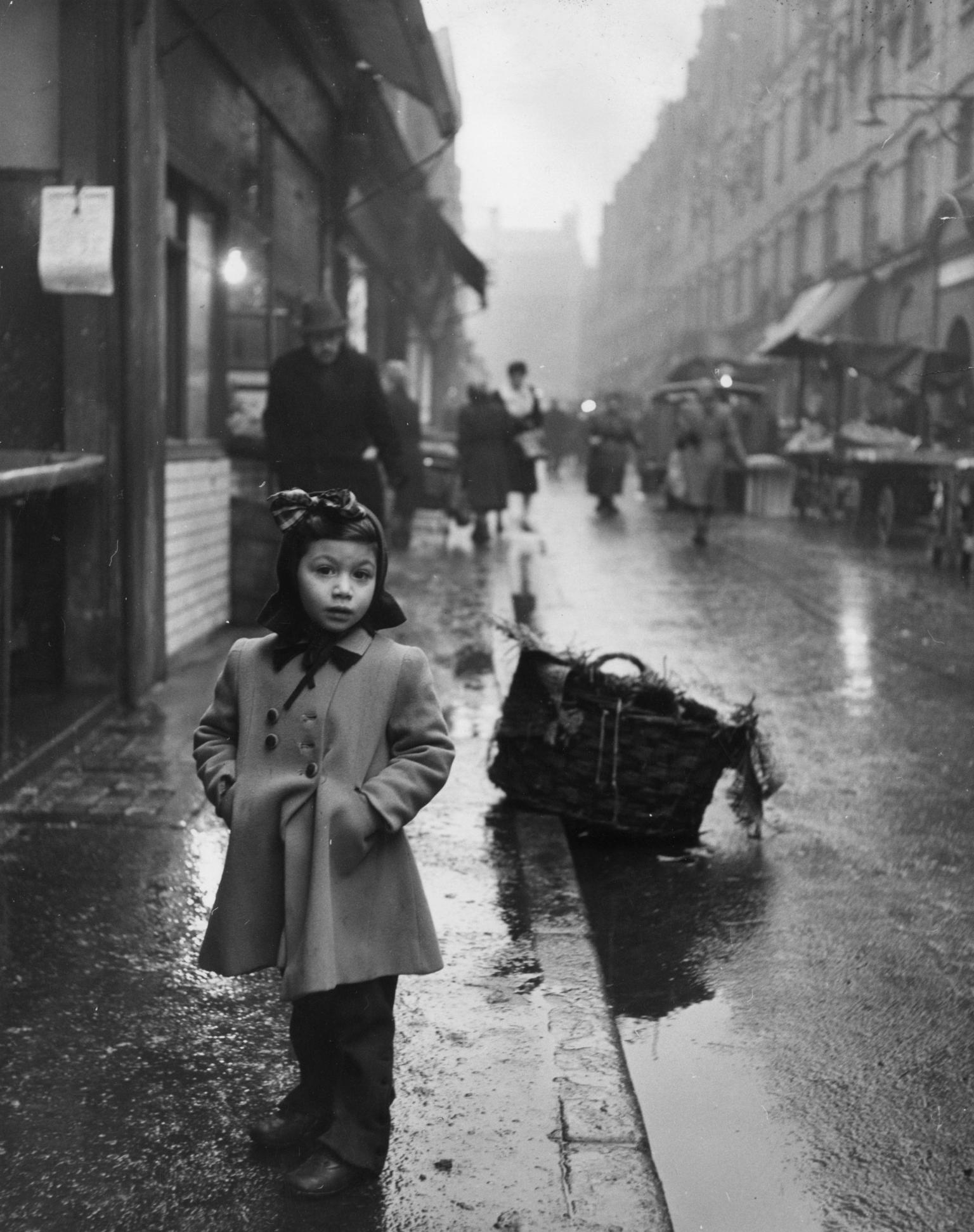 Photograph of little girl standing on rainy sidewalk of a busy city street wearing a coat and a bow facing the viewer, and basket on curb behind her.
