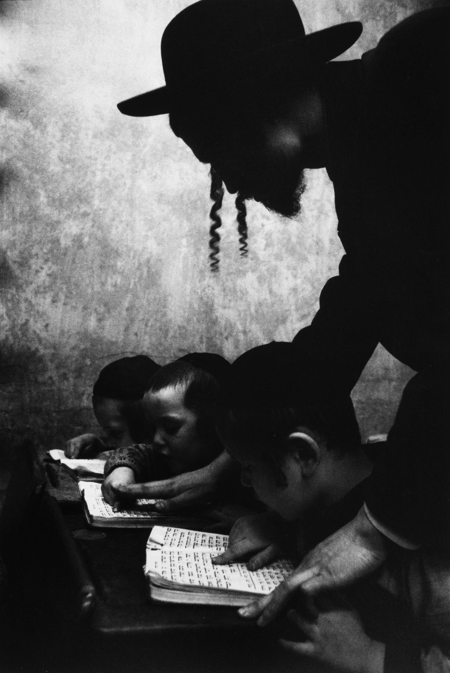 Photograph of man in a hat and sidelocks bending over a desk where three boys are reading Hebrew. 