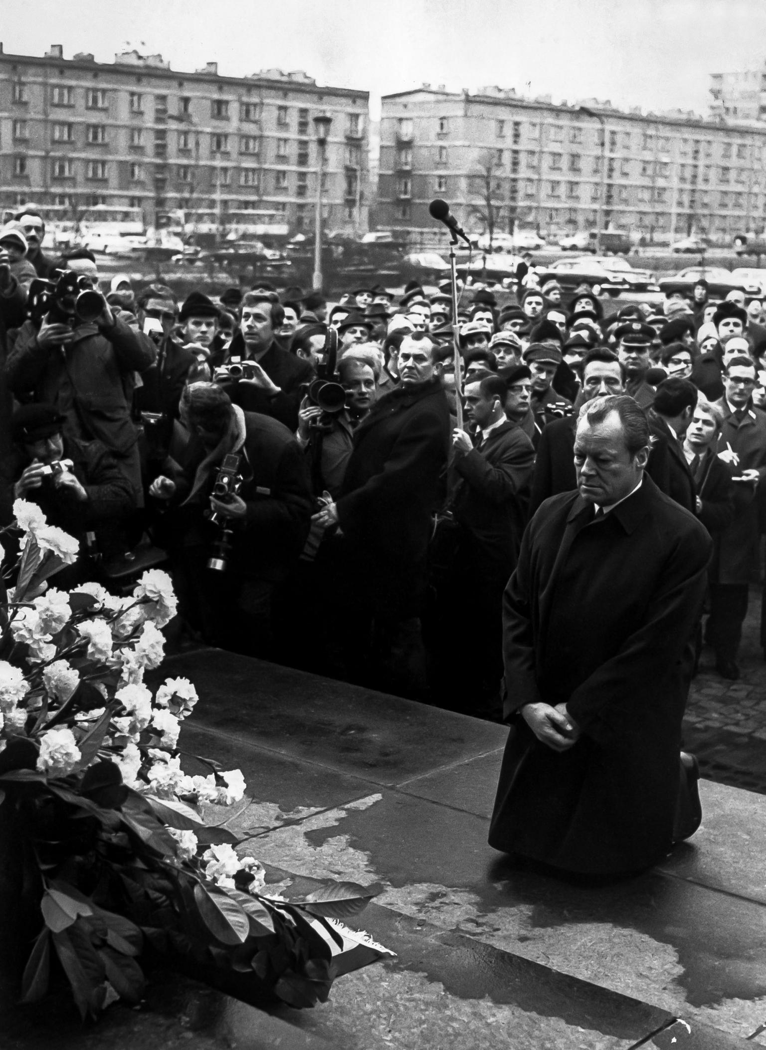 Photograph of a man in a long coat kneeling in front of a memorial wreath on a pedestal as crowd watches and photographers take photos.