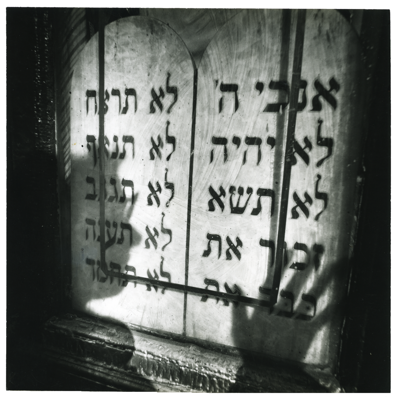 Photograph featuring shadow of a face and a hand over a wall with Hebrew text. 
