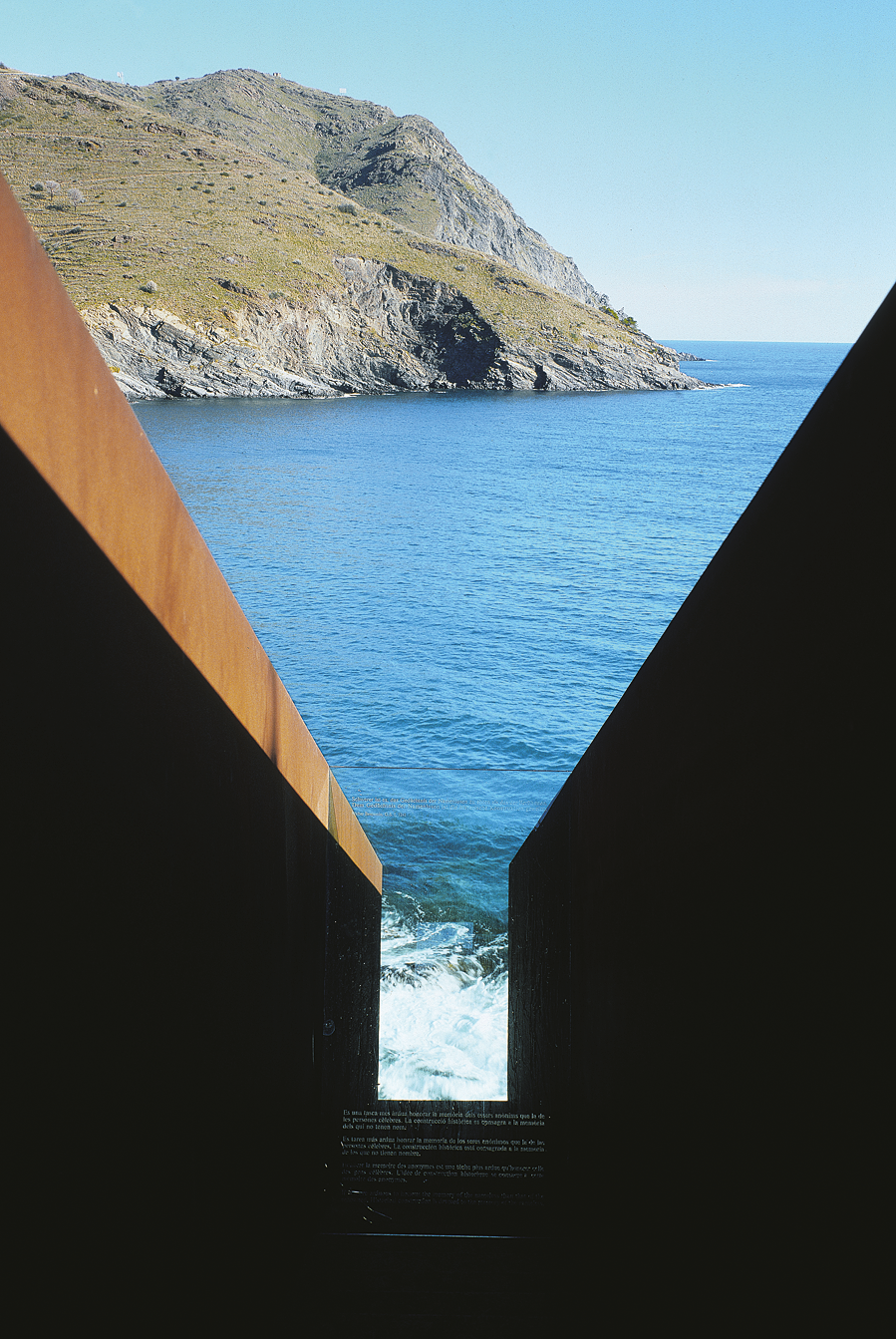 Photograph of metal sculpture in the shape of a V through which the ocean and rocky outcropping beyond is visible. 