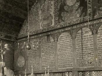 Interior of building of back wall with decorative images on top, Hebrew text in the middle, and lattice below, platform with railing in left foreground, and several podiums along wall.