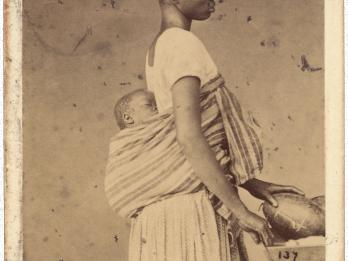 Full body profile photograph of person in profile with baby in shawl on her back.