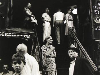 Photograph of people standing outside along stairwell and storefronts.