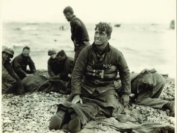 Photograph of soldiers on the beach, with man in center facing the viewer and looking up, as he kneels on top of a prone soldier.