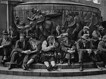 Photograph of men, women, and children seated on the steps of a large public monument with nude classical figures and a winged horse atop a circular base.