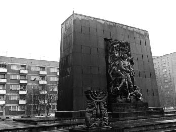 Photograph of tall, rectangular monument in front of apartment buildings, depicting seven figures gathered around central figure and smaller sculpture of a seven-branched menorah flanked by lions in front of monument in left foreground. 