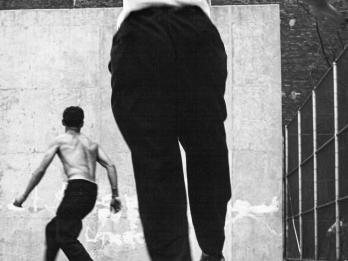 Photograph of two men playing handball, both of whom are turned away from the viewer and facing the wall. 